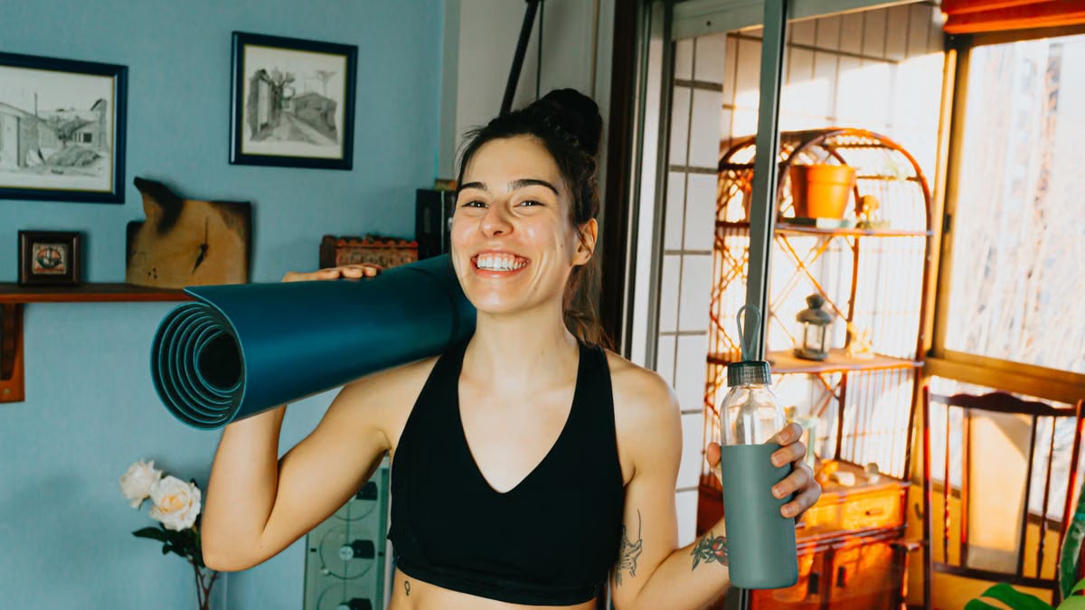 Cheerful woman with yoga mat and bottle ready for home workout in cozy room.